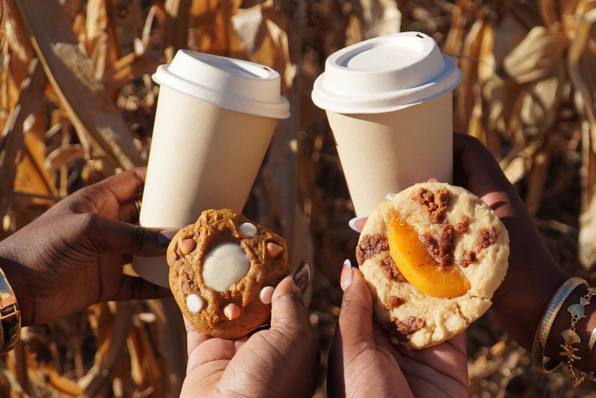 Two hands holding cookies and coffee cups against a cornfield background