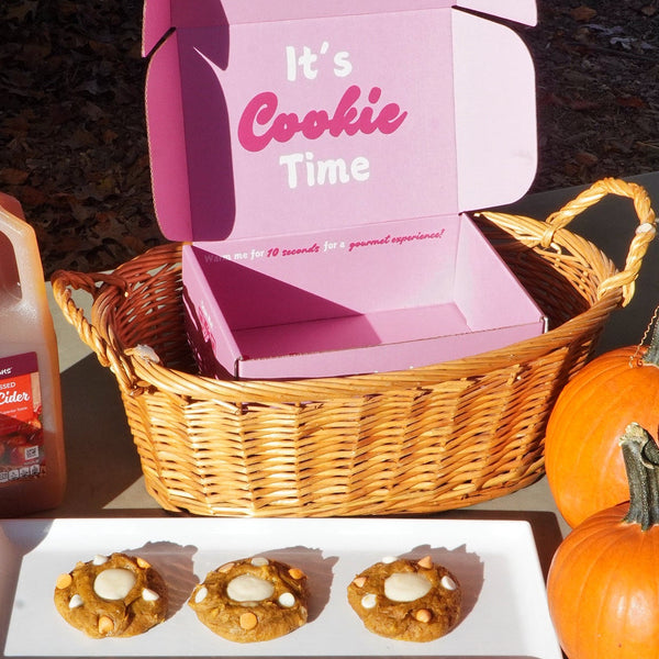 Cookie box labeled 'It's Cookie Time', cookies, orange juice, and pumpkins on a table outdoors.