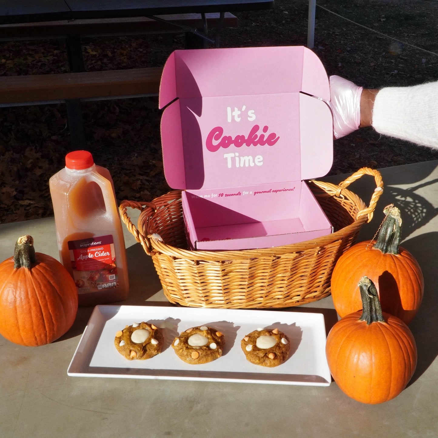 Pink cookie box with 'It's Cookie Time' text, cookies, pumpkins, and a basket on a table.