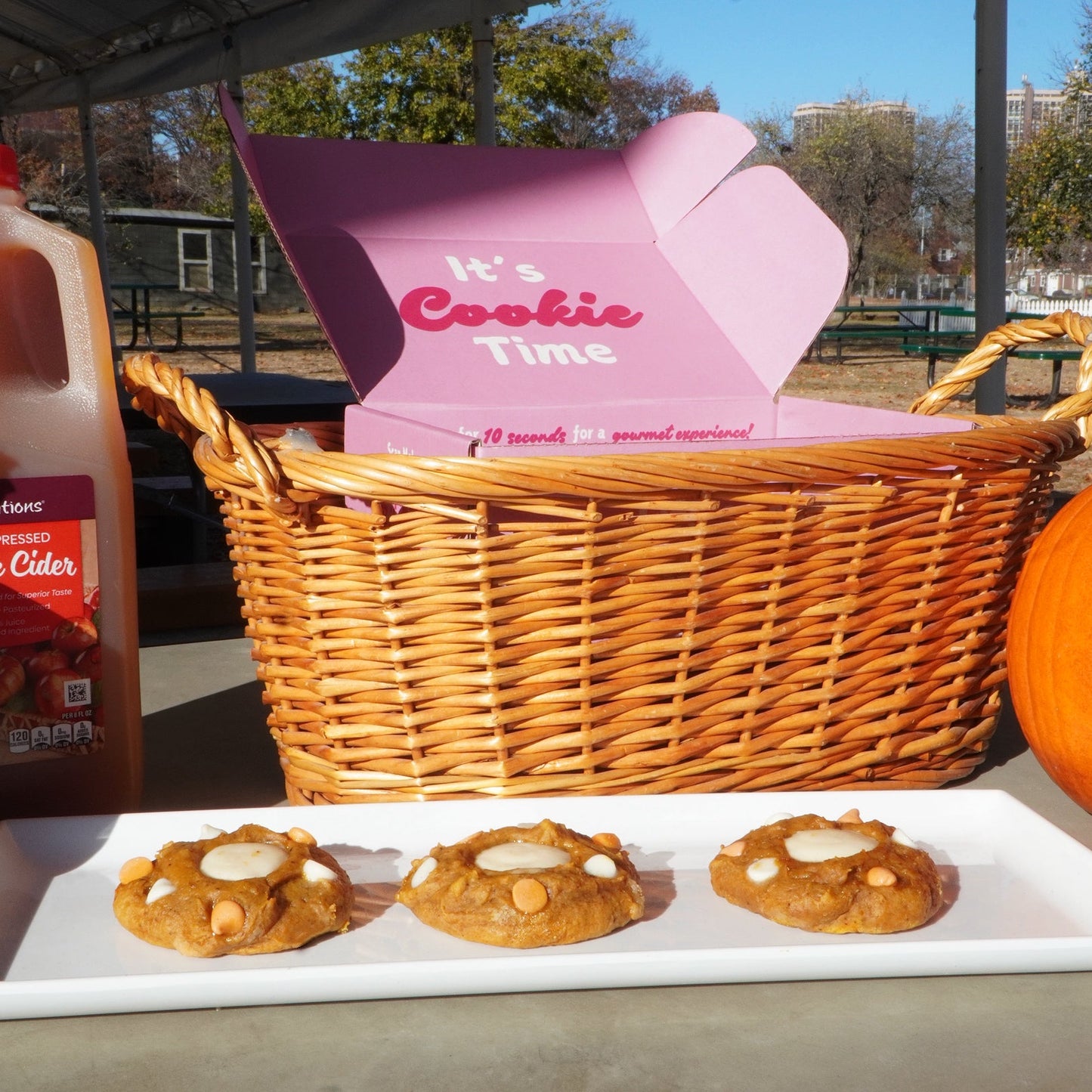 Basket with 'It's Cookie Time' box, cookies on a plate, and a bottle of apple cider outdoors.